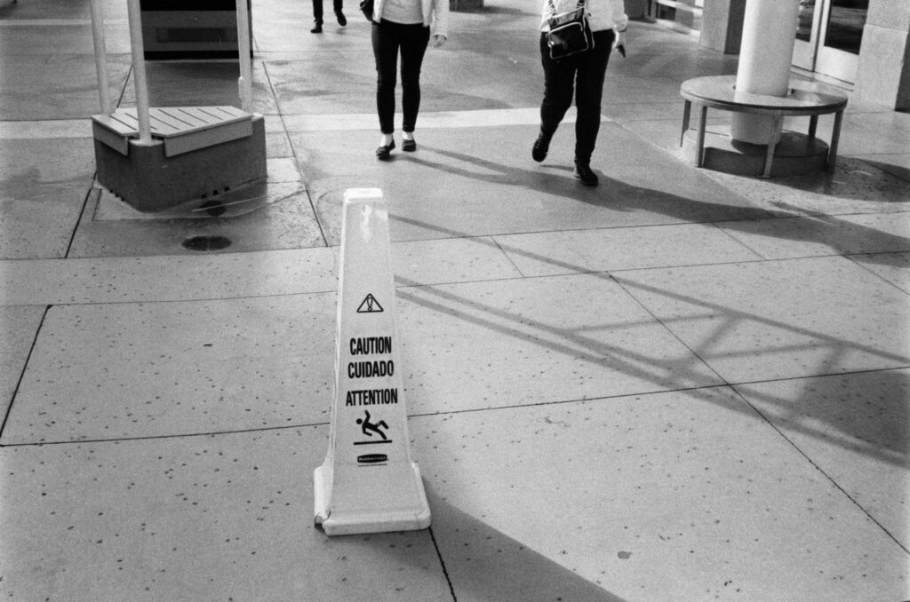 Yellow wet floor caution sign on an outdoor paved sidewalk near a building entrance with people walking in the background.